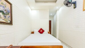 A narrow hotel room with white brick walls, a wooden bed, a red pillow, and a wall-mounted fan. - OYO 604 Queen Hotel (Hanoi)