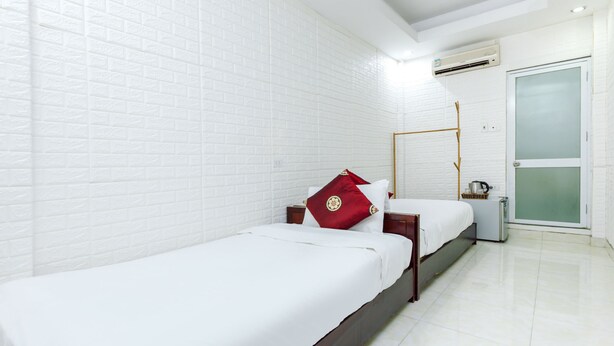 A hotel room with two beds, white brick walls, a white tiled floor, a wooden nightstand, a red pillow, a white bedspread, a wooden headboard, a small table with a kettle, and a sliding glass door. - OYO 604 Queen Hotel (Hanoi)