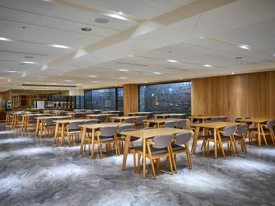 A spacious dining area with wooden tables and chairs, large windows, and a modern ceiling design.