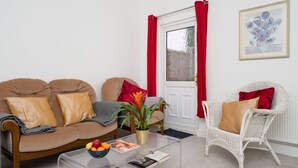 Een woonkamer met een bruine bank, een witte rieten stoel, een glazen salontafel met tijdschriften en een schaal fruit, en een raam met rode gordijnen.