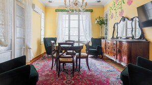 Uma sala de jantar com mesa redonda, cadeiras, lustre, espelho grande e decoração floral na parede.