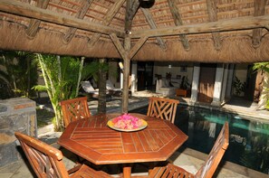 An outdoor dining area with a round wooden table and chairs, a thatched roof, and a pool in the background.