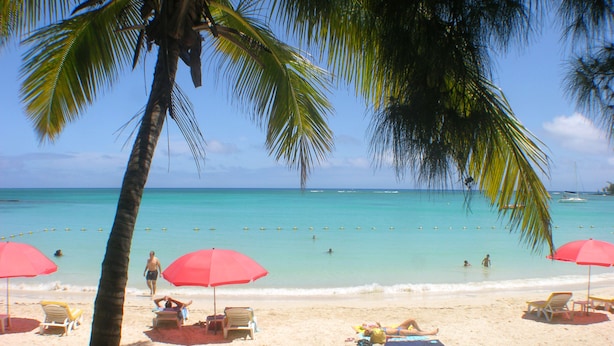 A beach with palm trees, clear turquoise water, and people sunbathing under red umbrellas. - Villa Oasis 1 no 7 (Grand-Baie)