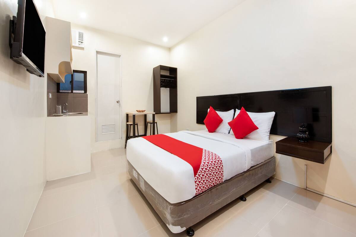 A modern hotel room with a bed, bedside table, TV, and a small kitchenette area.