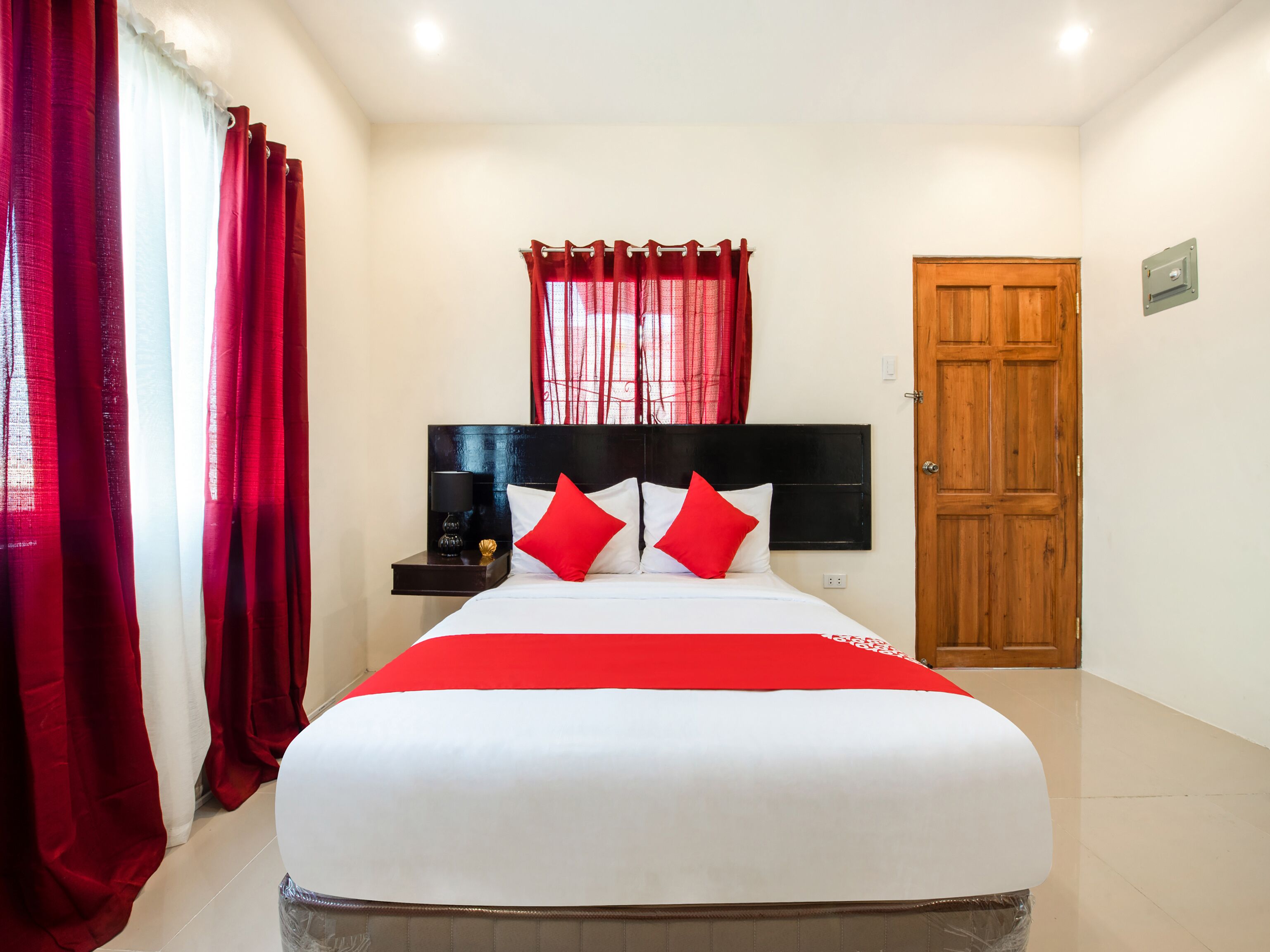A hotel room with a large bed, red curtains, a wooden door, and a nightstand with a lamp.