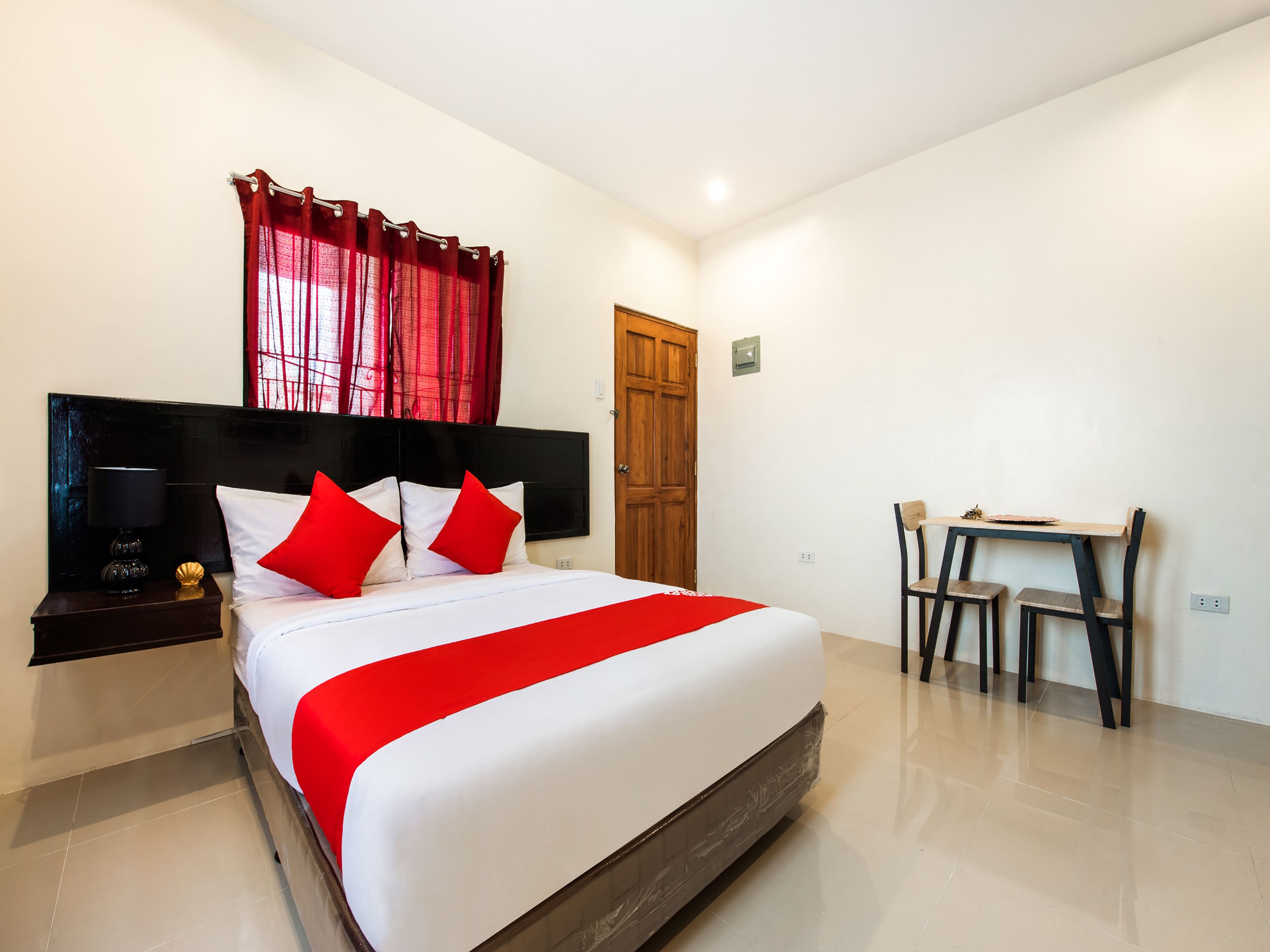 A hotel room with a large bed, a wooden headboard, a desk with two chairs, and a red curtain.