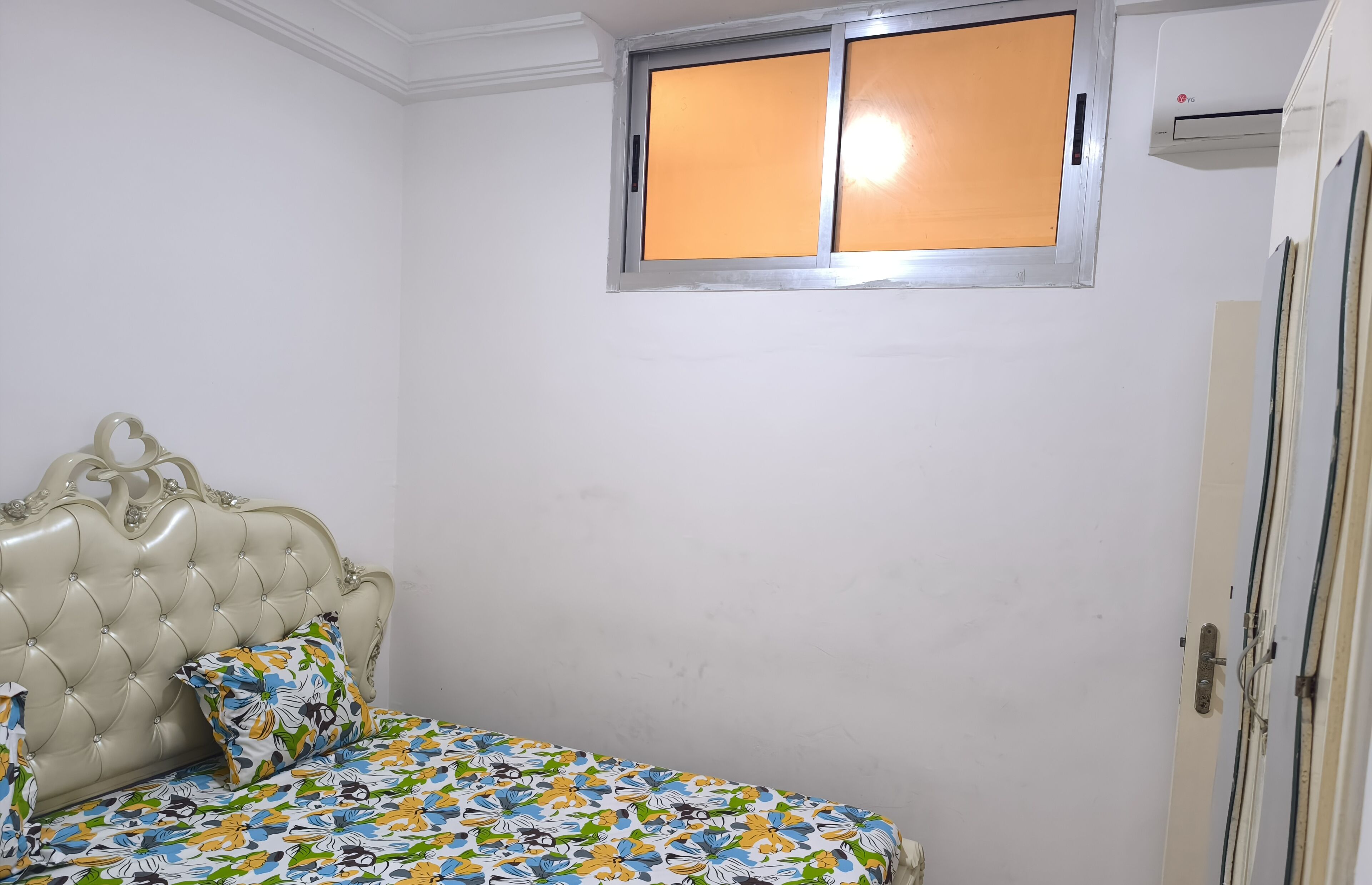 A bedroom with a tufted headboard, a floral bedspread, a window with a metal frame, and an air conditioning unit.