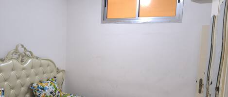 Une chambre à coucher avec une tête de lit capitonnée, une couvre-lit à motifs floraux, une fenêtre avec un cadre en métal et un appareil de climatisation.