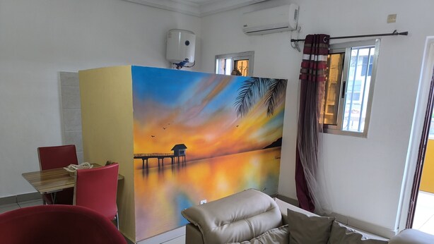 A room with a large painting of a sunset over water, a red chair, a beige sofa, and a small table with a newspaper. - Résidence Gian (Libreville)
