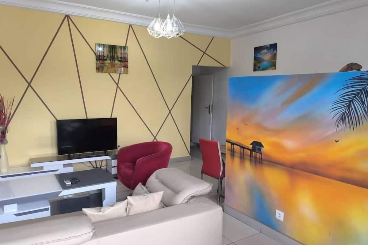 Uma sala de estar com televisão, uma cadeira vermelha, um sofá bege e um quadro grande na parede.