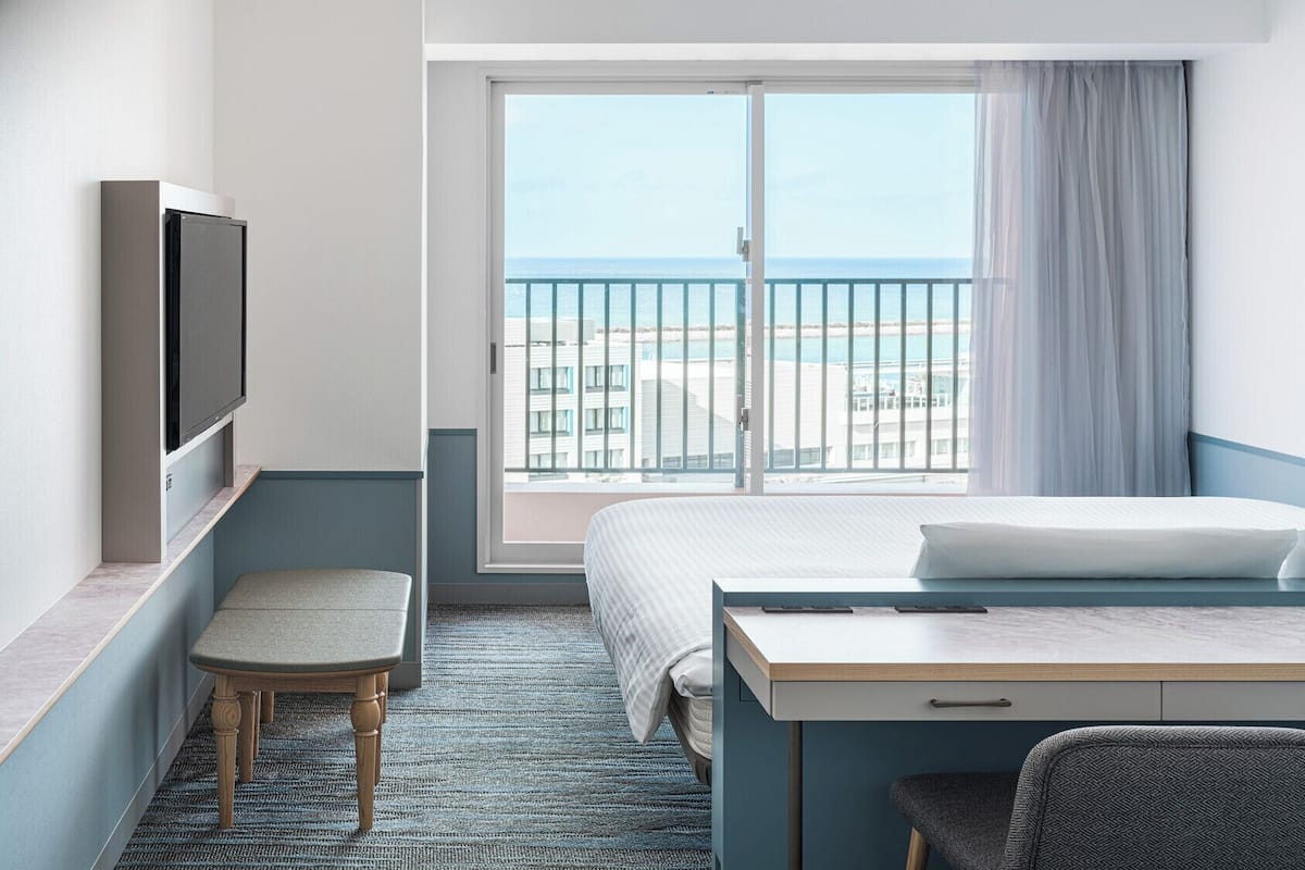A hotel room with a bed, a desk, a chair, a TV, and a large window with a view of the beach and ocean.