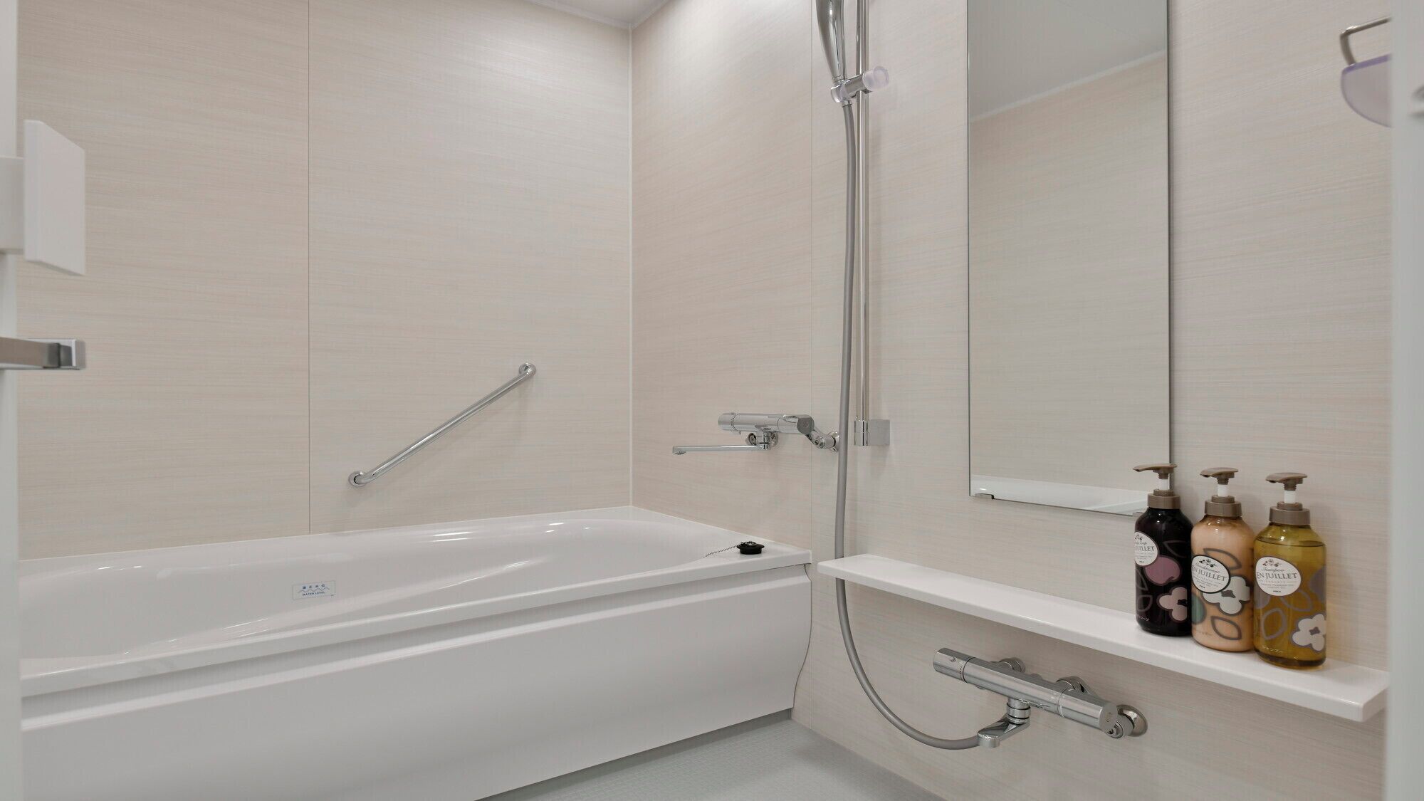 A modern bathroom with a white bathtub, a glass shower enclosure, a handrail, a towel rack, and a shelf with toiletry bottles.
