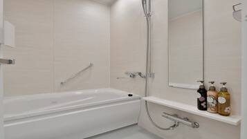 A modern bathroom with a white bathtub, a glass shower enclosure, a handrail, a towel rack, and a shelf with toiletry bottles.
