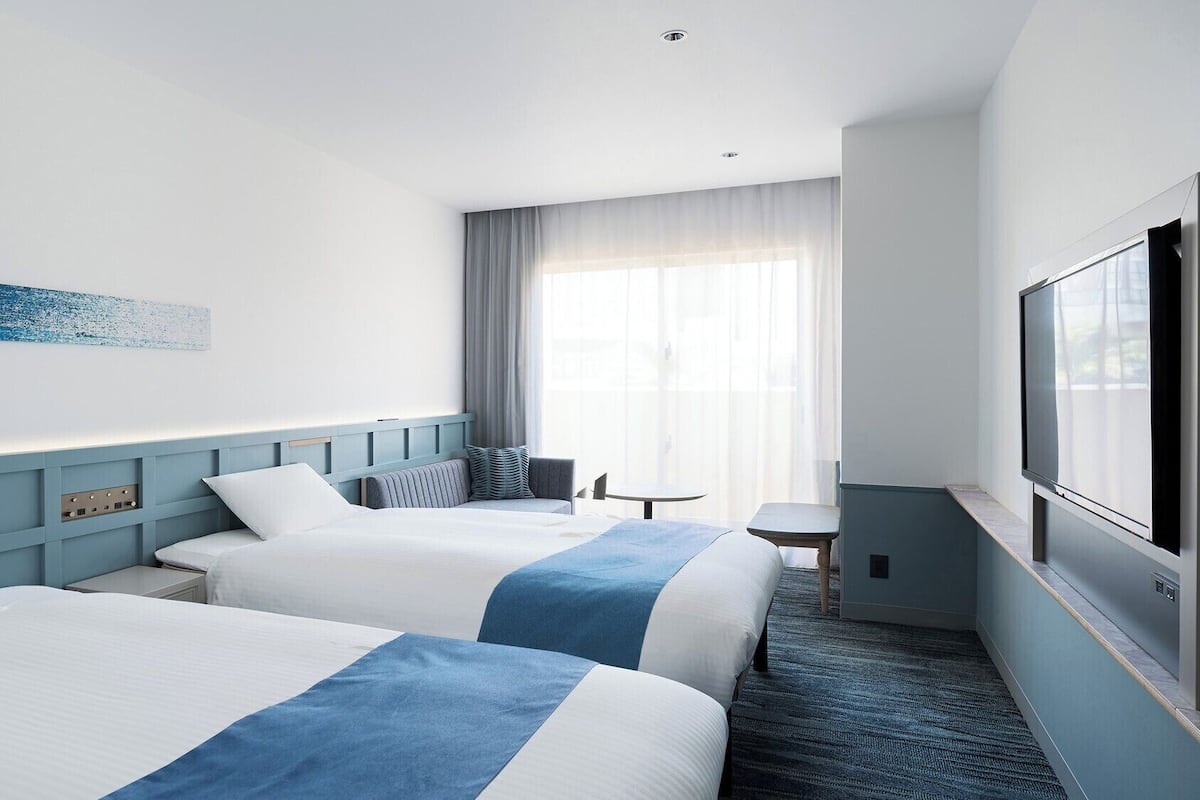 A hotel room with two beds, a TV, a small table, and a window with curtains.