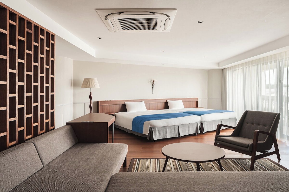 A hotel room with a bed, two bedside tables, a sofa, a chair, a TV, and a large window with curtains.