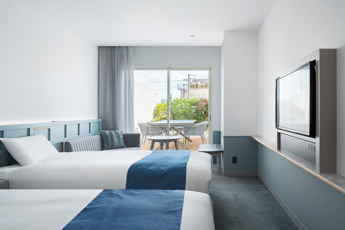 A hotel room with two beds, a TV, a balcony with a table and chairs, and a painting on the wall.