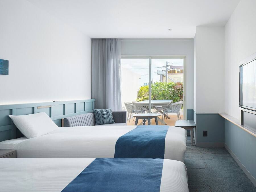 Habitación de hotel con dos camas, televisión, balcón con mesa y sillas, y un cuadro en la pared.