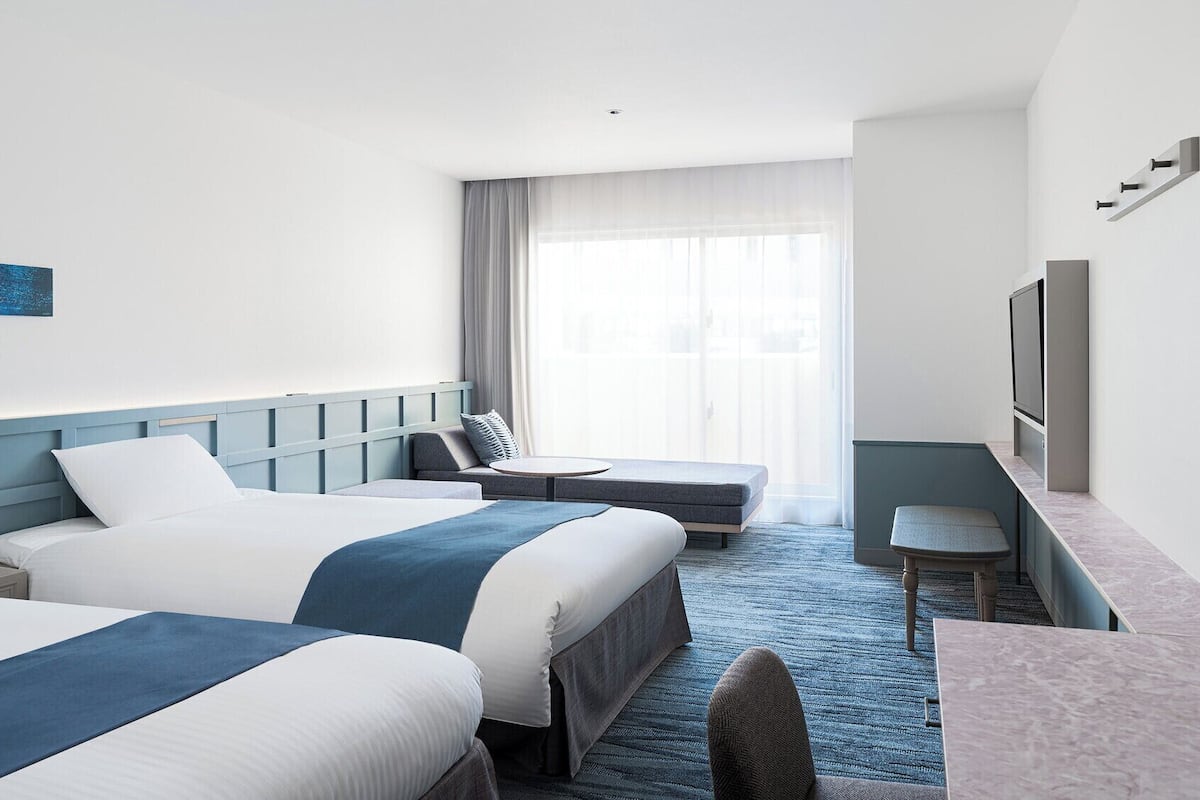 A hotel room with two beds, a desk, a chair, and a TV.