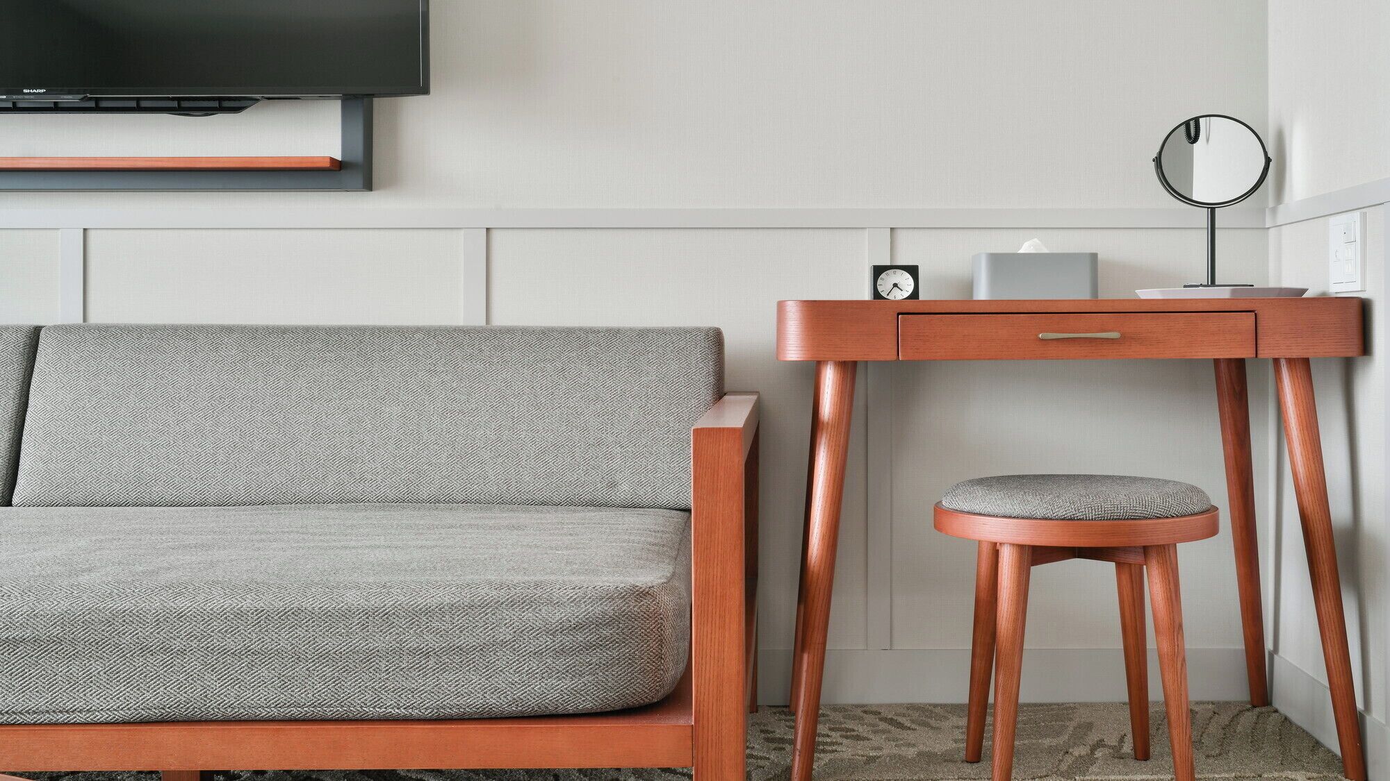 Una habitación moderna con un sofá gris, un escritorio de madera con una silla, un espejo redondo y un reloj.