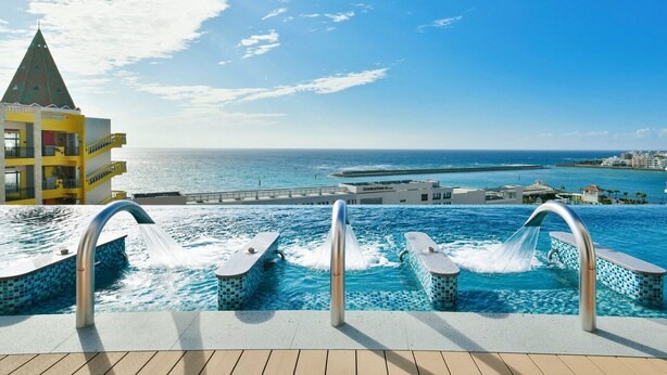 Zona de piscina con tres tableros de clavados, vista al océano y un edificio con balcones.