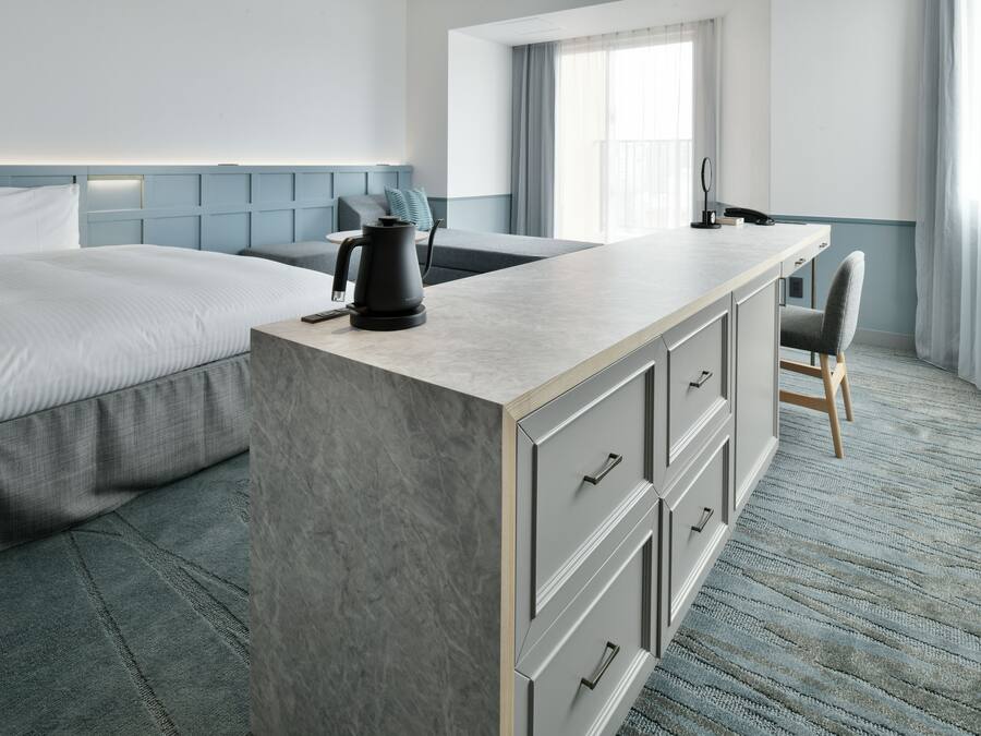 A hotel room with a bed, a small desk with a chair, a coffee maker, and a view of the outdoors.