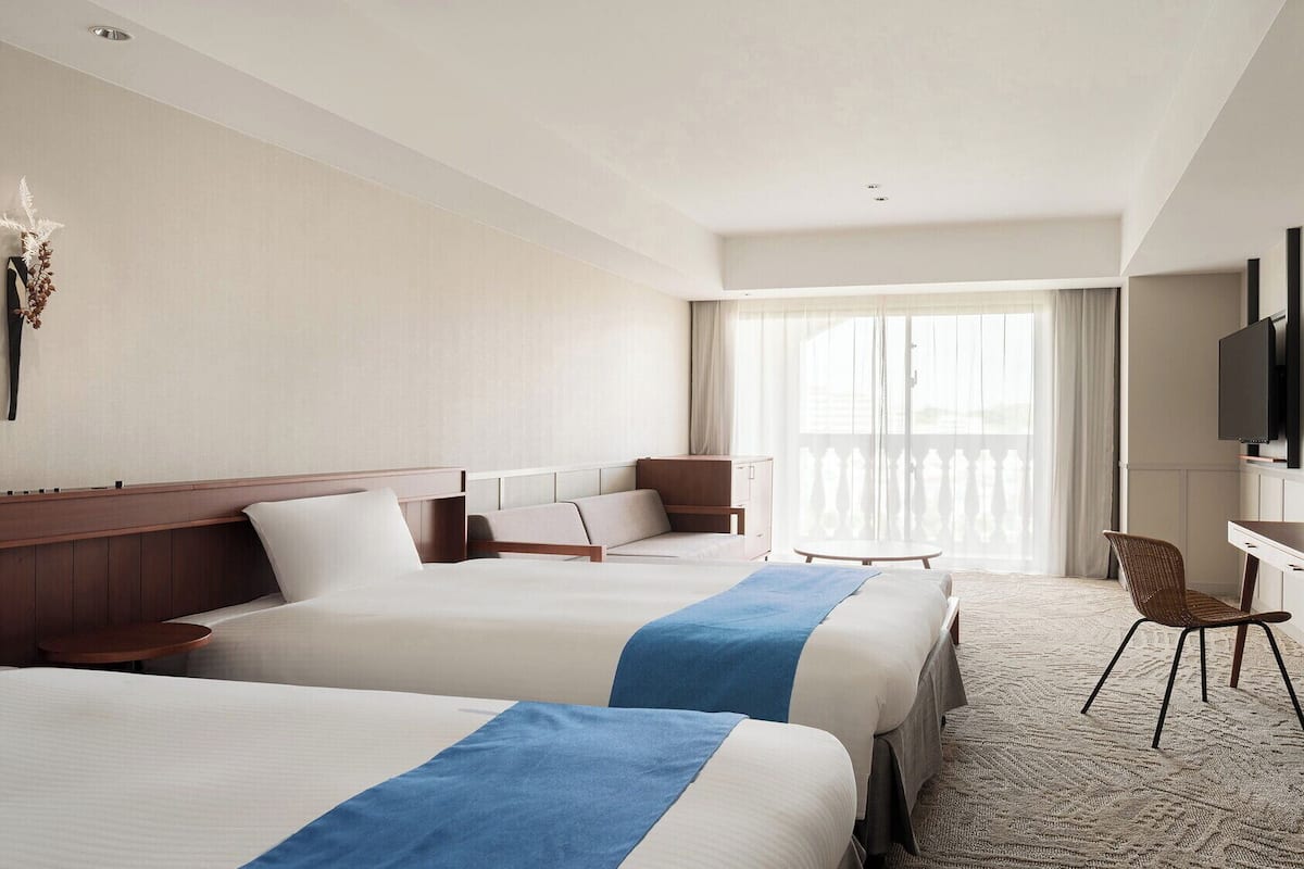 A hotel room with two beds, a desk, a chair, a television, and a large window with curtains.