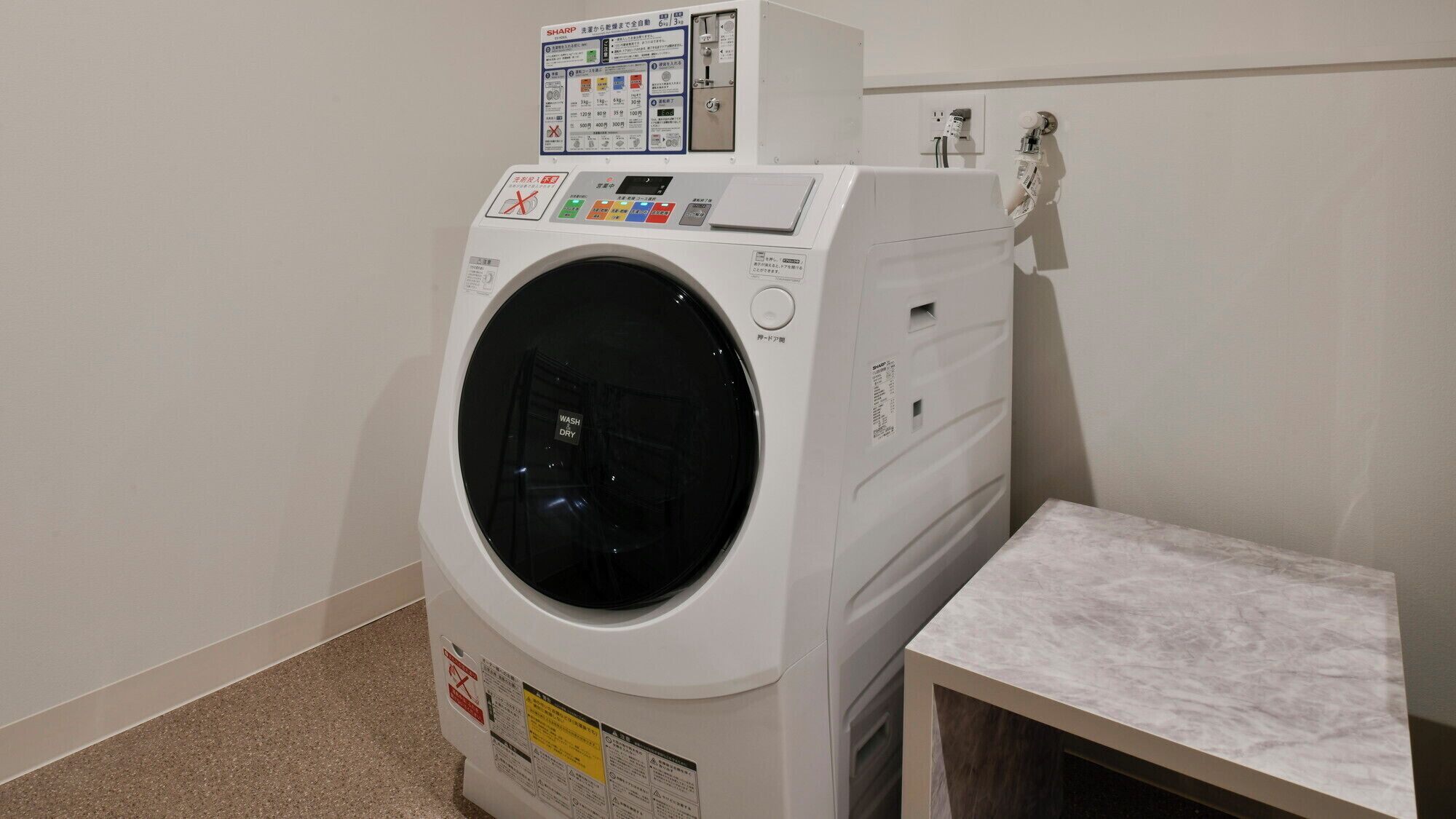 A front-loading washing machine with a control panel, a small table, and a wall-mounted electrical outlet.