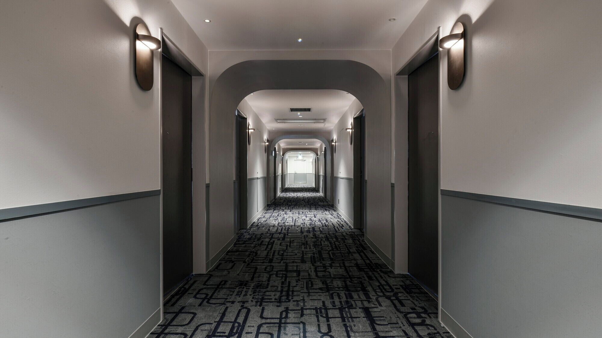 A long, narrow hallway with a patterned carpet, walls with light fixtures, and a ceiling with recessed lighting.
