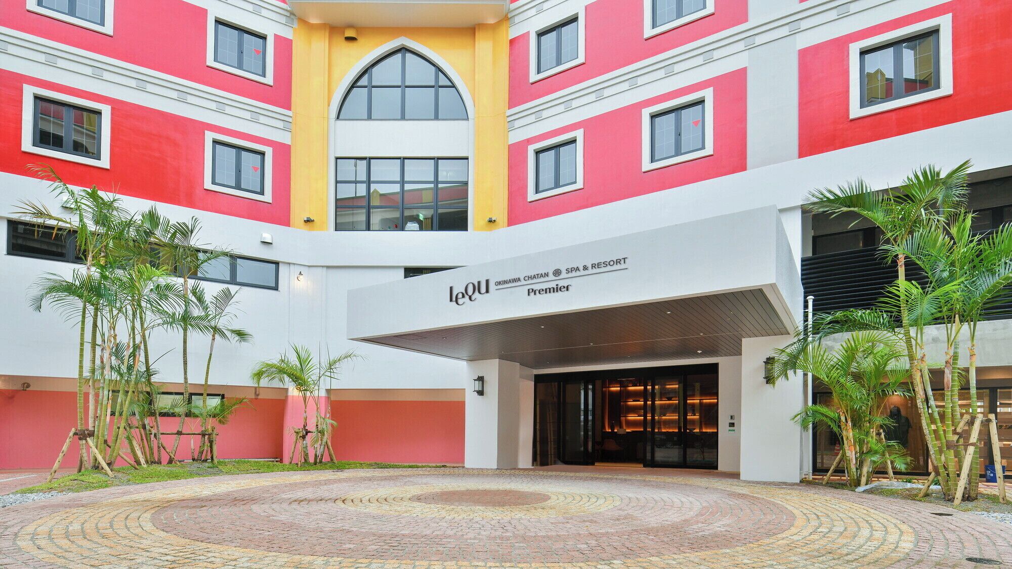 A hotel with a red and white facade, a central courtyard with palm trees, and a sign for 'LeQu Okinawa Chatan Spa & Resort Premier'.