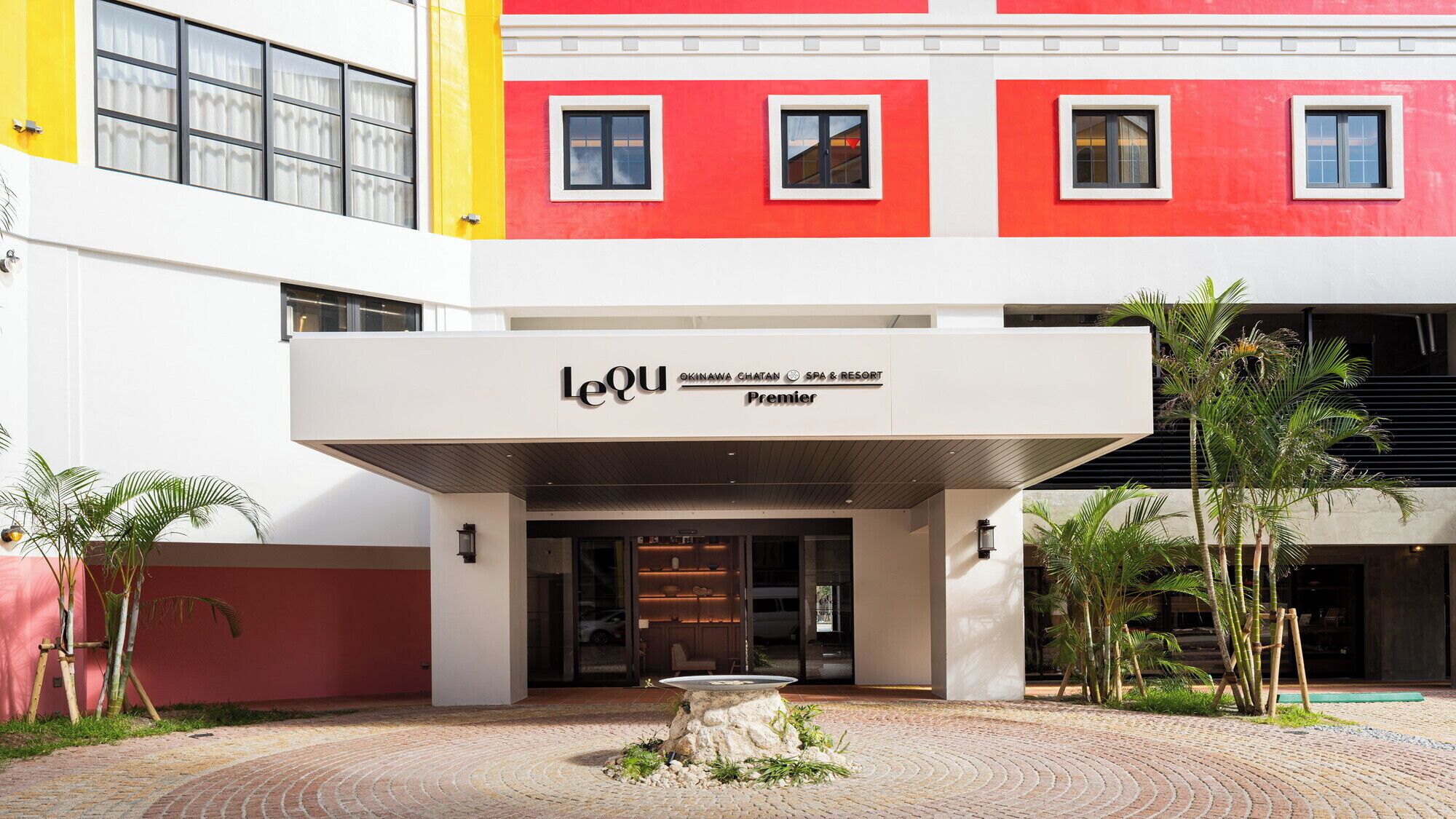 A building with a prominent sign for 'Lequ' hotel, featuring a modern architectural design with a red and white facade, a covered entrance, and a landscaped courtyard with palm trees.