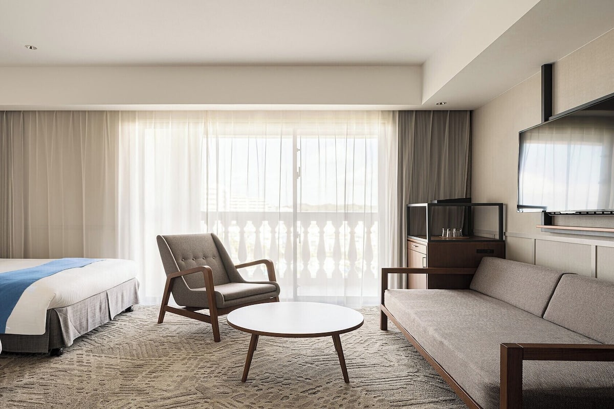 A hotel room with a bed, a chair, a sofa, a TV, and a large window with curtains.