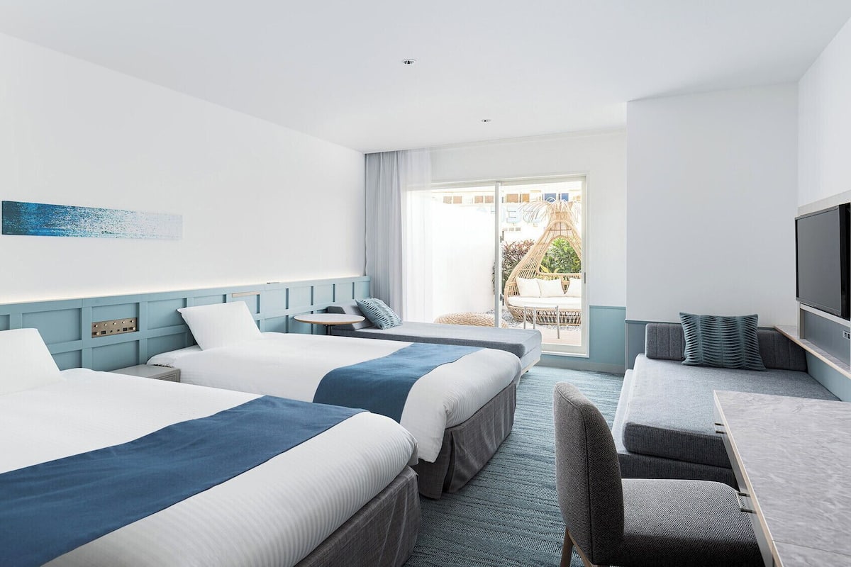 A hotel room with two beds, a desk, a chair, a TV, and a large window with a view of the outdoors.