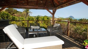 A covered outdoor seating area with wicker furniture and a glass table, overlooking a lush garden and a mountainous landscape. - Villa Acassia with 2 rooms (Black River)