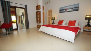 A bedroom with a large bed, a red chair, a painting of a sailboat, and a wooden shelf unit. - Villa Acassia with 2 rooms (Black River)