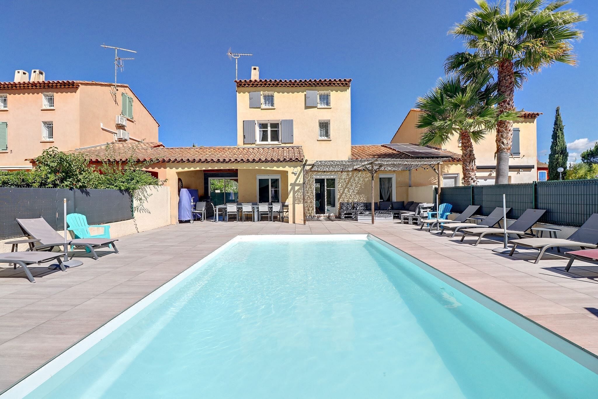 A swimming pool with lounge chairs and umbrellas, surrounded by buildings with balconies and a clear sky.