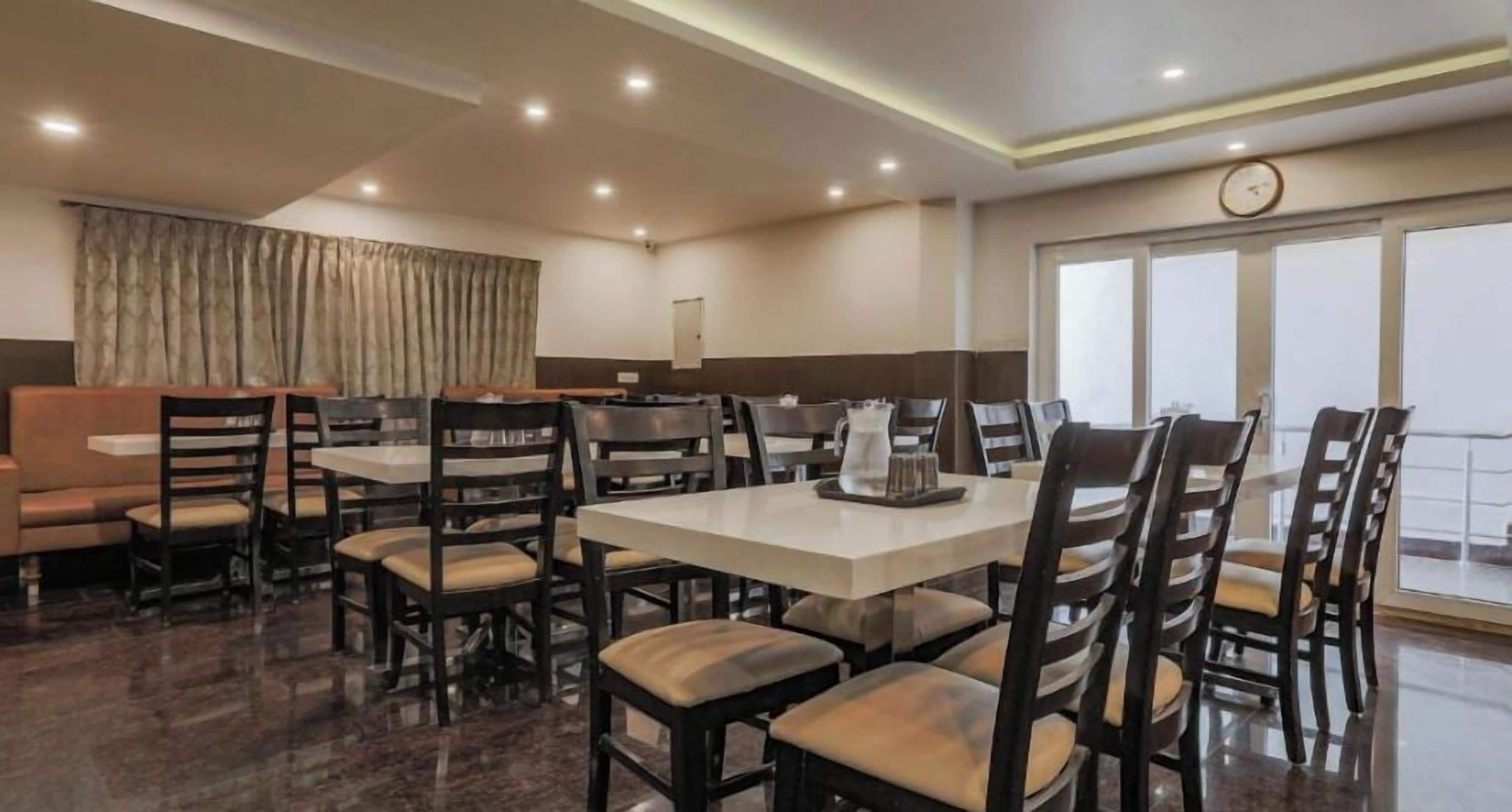 Comedor de hotel con varias mesas y sillas, un reloj en la pared y vistas al exterior a través de grandes ventanales.