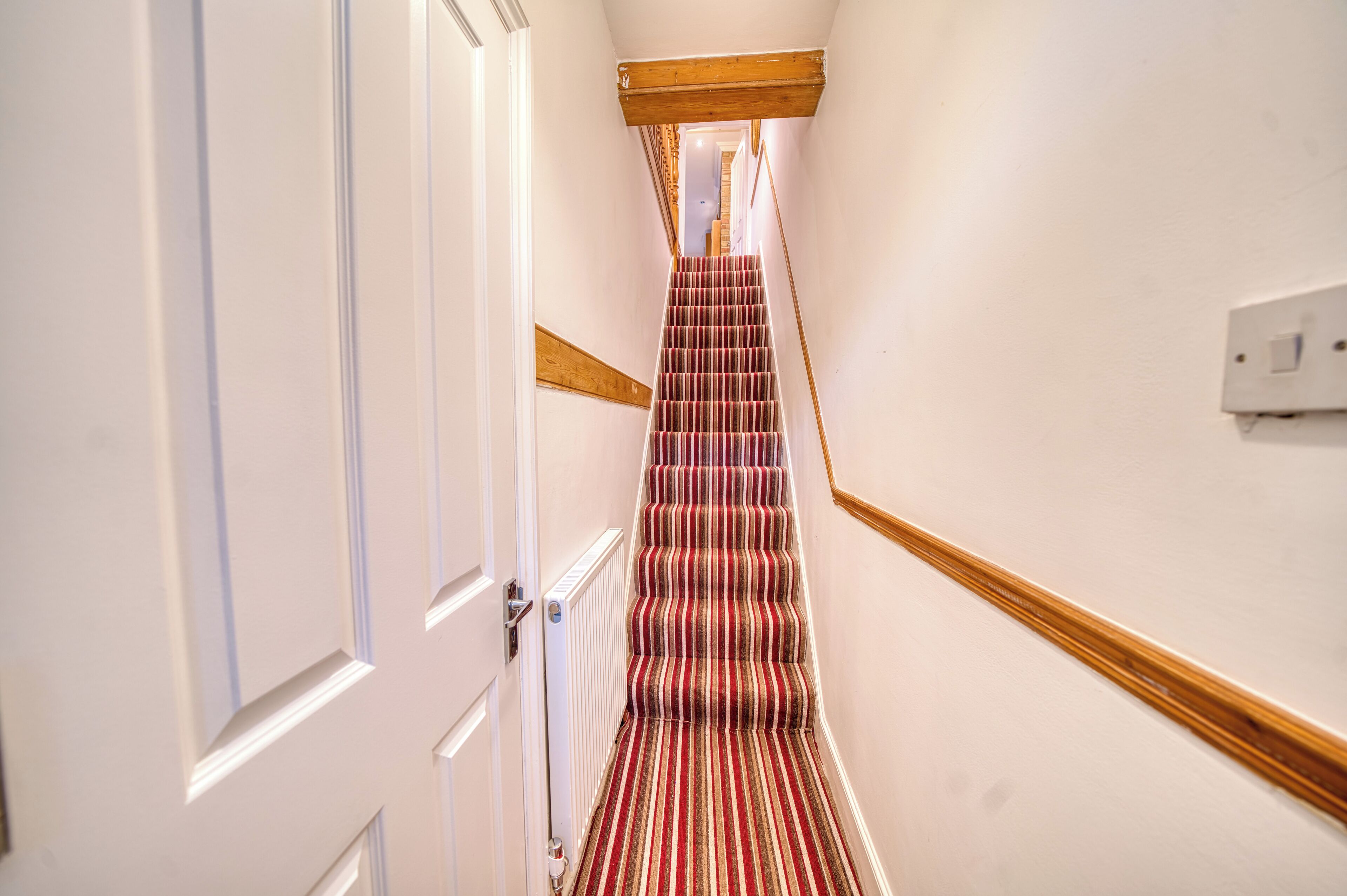 Un pasillo estrecho con barandilla de madera, alfombra a rayas roja y beige, y paredes blancas.