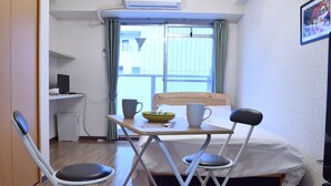 Una habitación pequeña y funcional con cama, una mesa con sillas, un escritorio con computadora y una ventana con cortinas.
