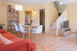 A living room with a red sofa, a dining table with chairs, a bookshelf, a TV on a stand, and a staircase.