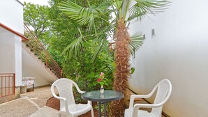 A small outdoor patio with two white plastic chairs and a round table, surrounded by lush greenery and a palm tree.