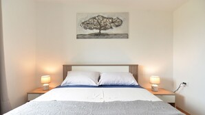 Cama bem arrumada com colcha branca e azul, duas almofadas brancas e duas luminárias de cabeceira em mesas de cabeceira de madeira.