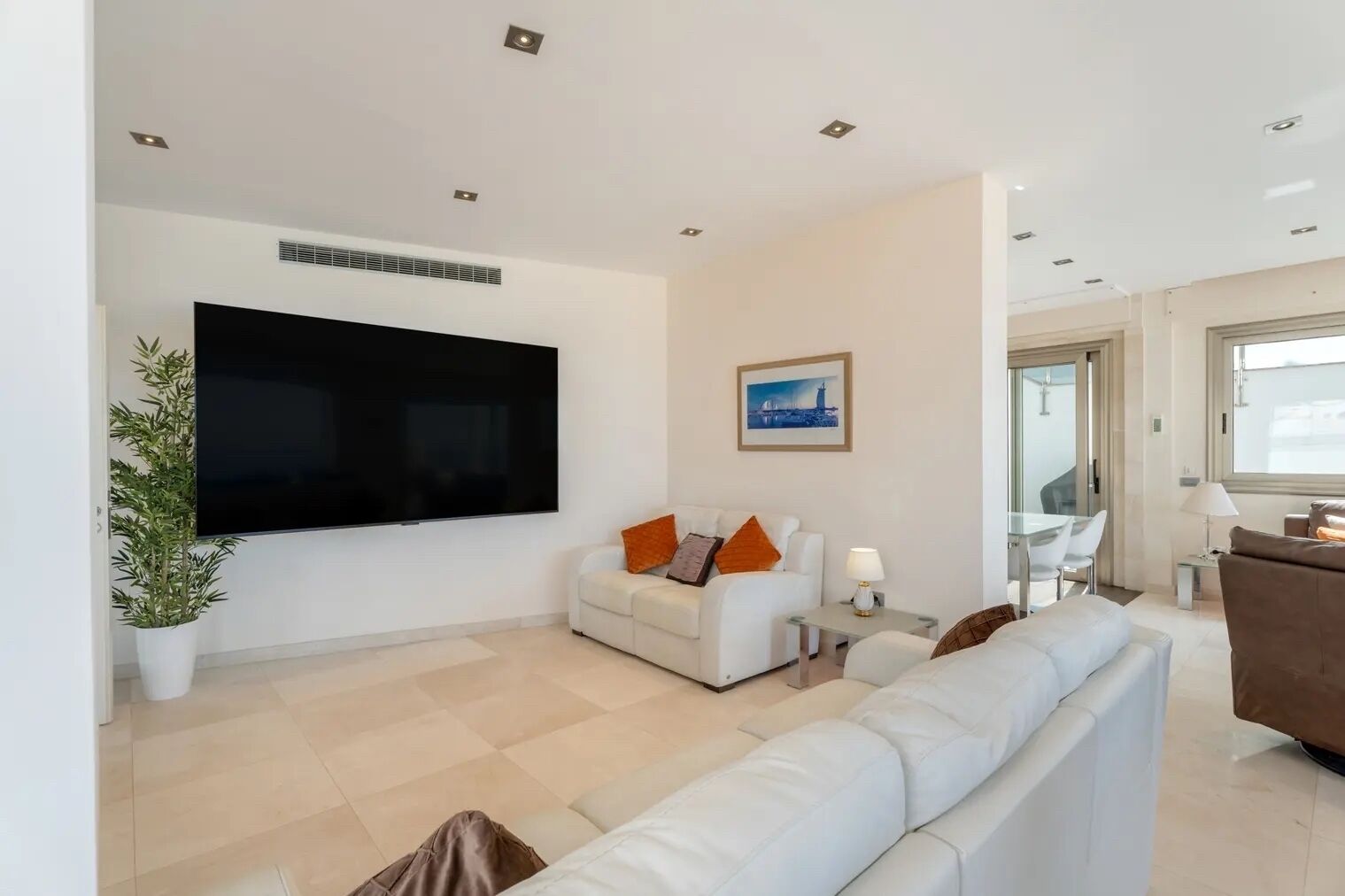 A modern living room with a flat-screen TV, a white sofa, a brown sofa, a dining area with a table and chairs, and a potted plant.