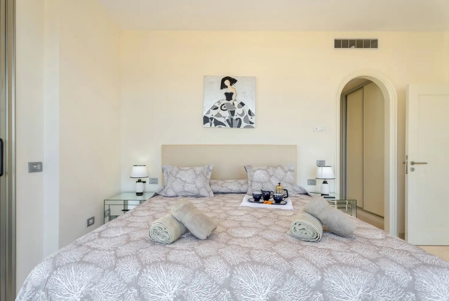 Une chambre avec un grand lit, deux tables de chevet avec des lampes, une œuvre d’art murale et une porte donnant sur une autre pièce.