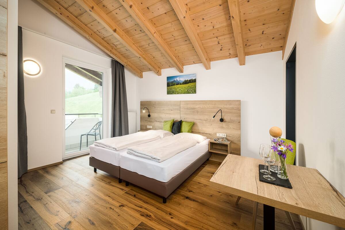 Un dormitorio con techo de madera, una cama con dos almohadas, una mesita de noche, una mesa con un florero y una ventana con vistas a un campo verde.