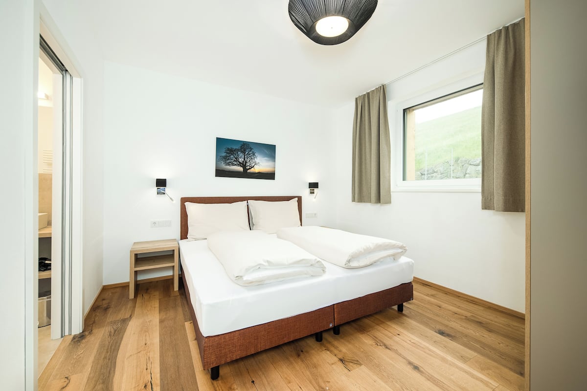 Un dormitorio con una cama de madera, una mesita de noche, una lámpara de pared y una ventana con cortinas.