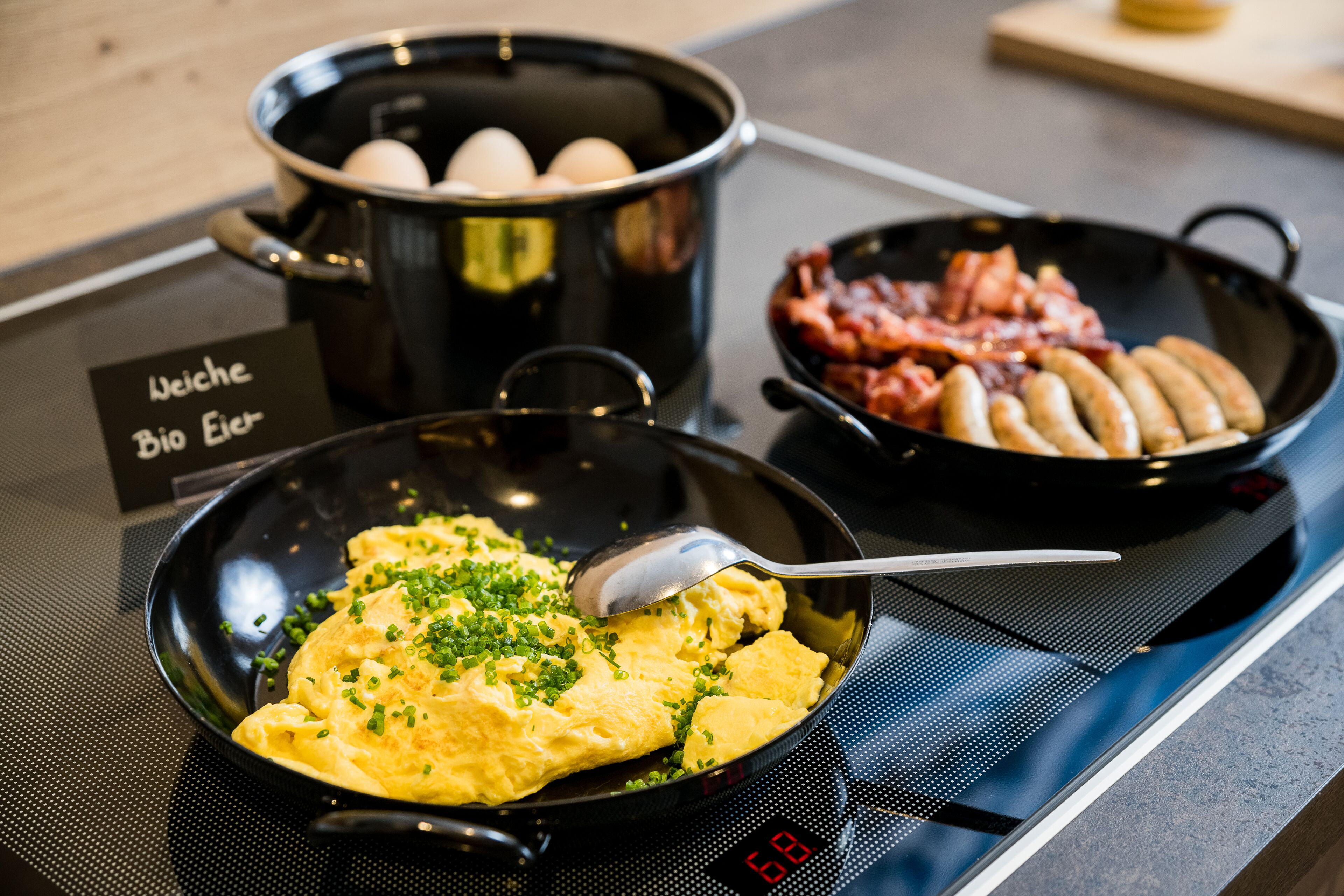 A kitchen counter with a frying pan containing scrambled eggs garnished with chives, a pot with eggs, and a pan with bacon and sausages.