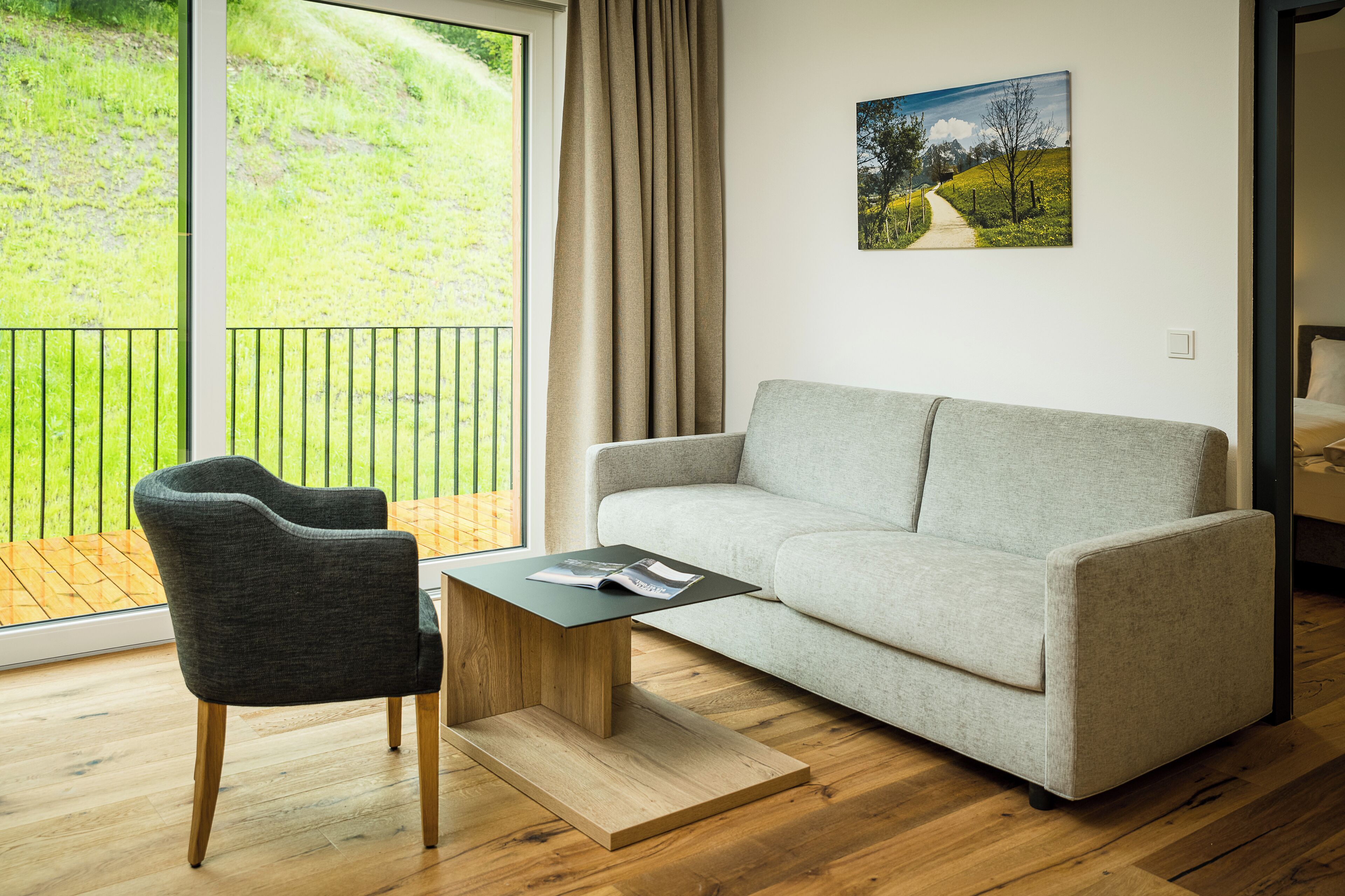 A modern living room with a grey sofa, a dark grey armchair, and a wooden coffee table with a magazine. A painting of a landscape hangs on the wall, and a balcony with a view of greenery is visible.