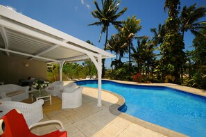 A pool area with white wicker furniture, a covered seating area, and palm trees in the background. - Villa Philibert (Surinam)