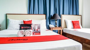 A hotel room with two beds, each with white bedding and red pillows. There is a red blanket on the bed, a lamp on a bedside table, and a RedDoorz promotional package on the bed. - RedDoorz Plus near Rizal Junction (Puerto Princesa)
