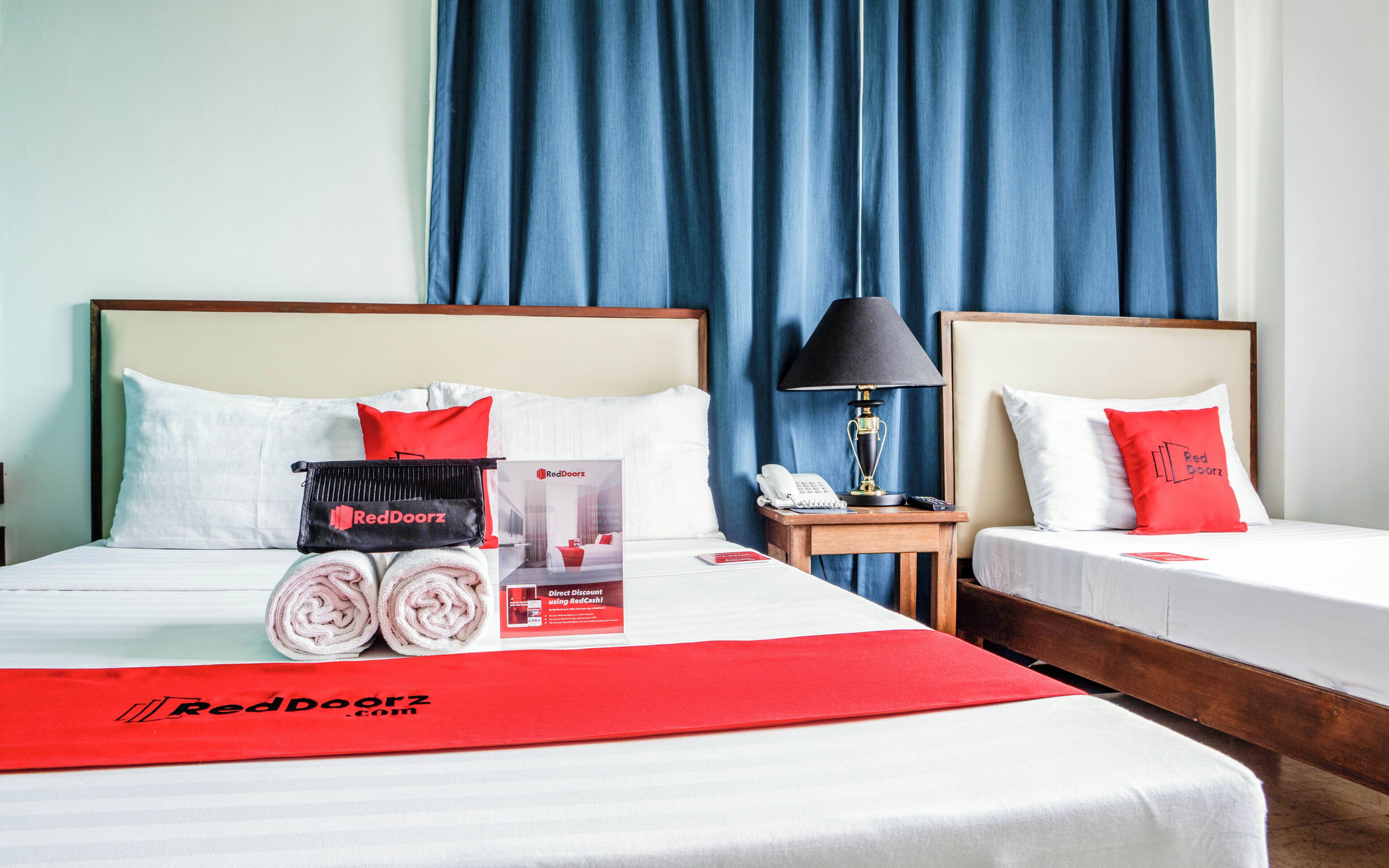 A hotel room with two beds, each with white bedding and red pillows. There is a red blanket on the bed, a lamp on a bedside table, and a RedDoorz promotional package on the bed.